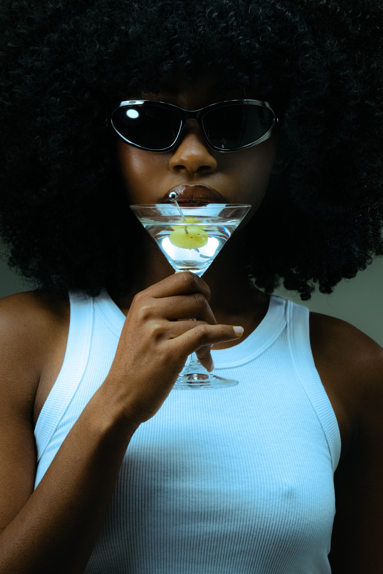 Portrait of a woman with dark curly hair and black sunglasses holding a martini glass against a dark background