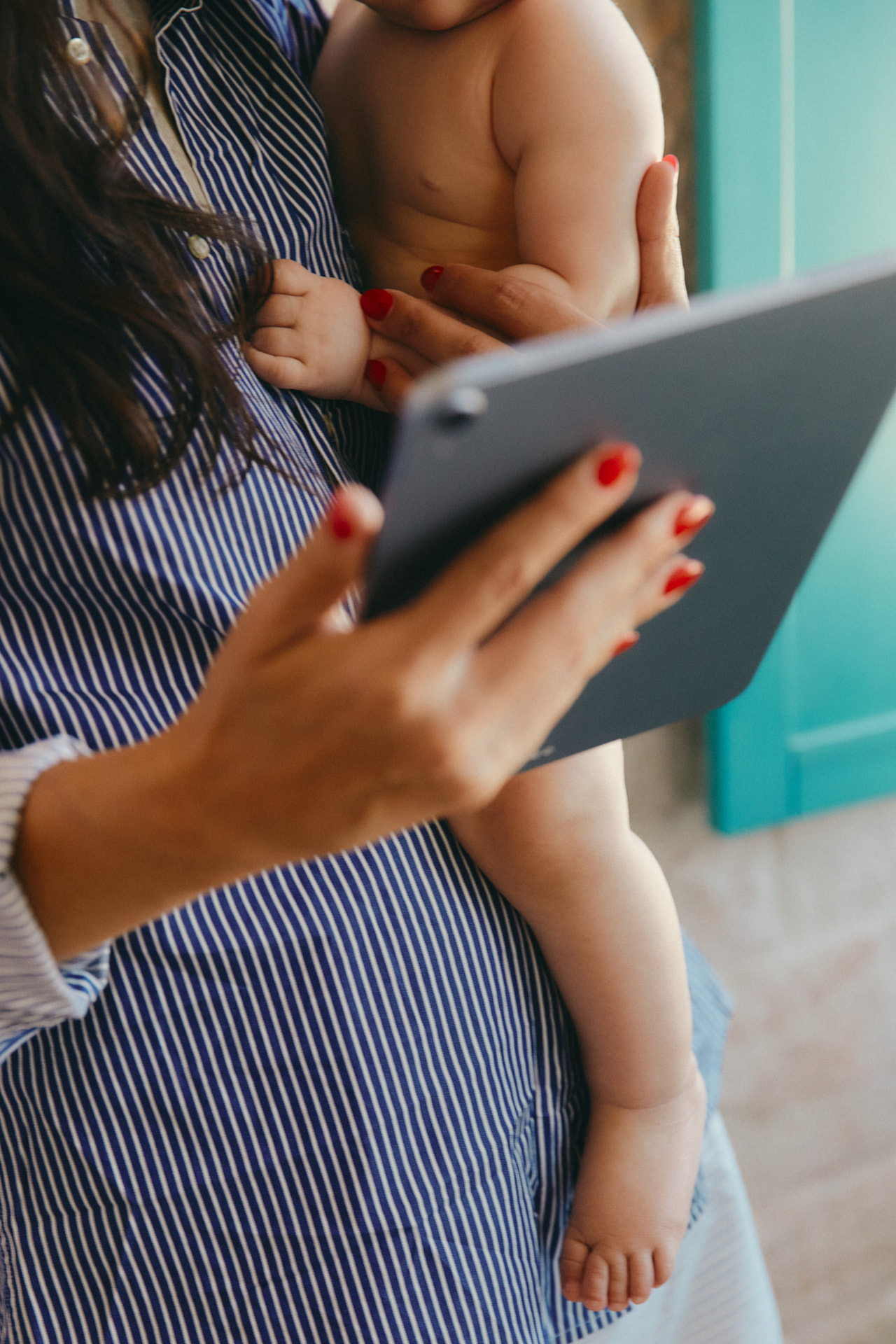 Mother in a blue-and-white striped shirt holding her baby against her body while using a tablet