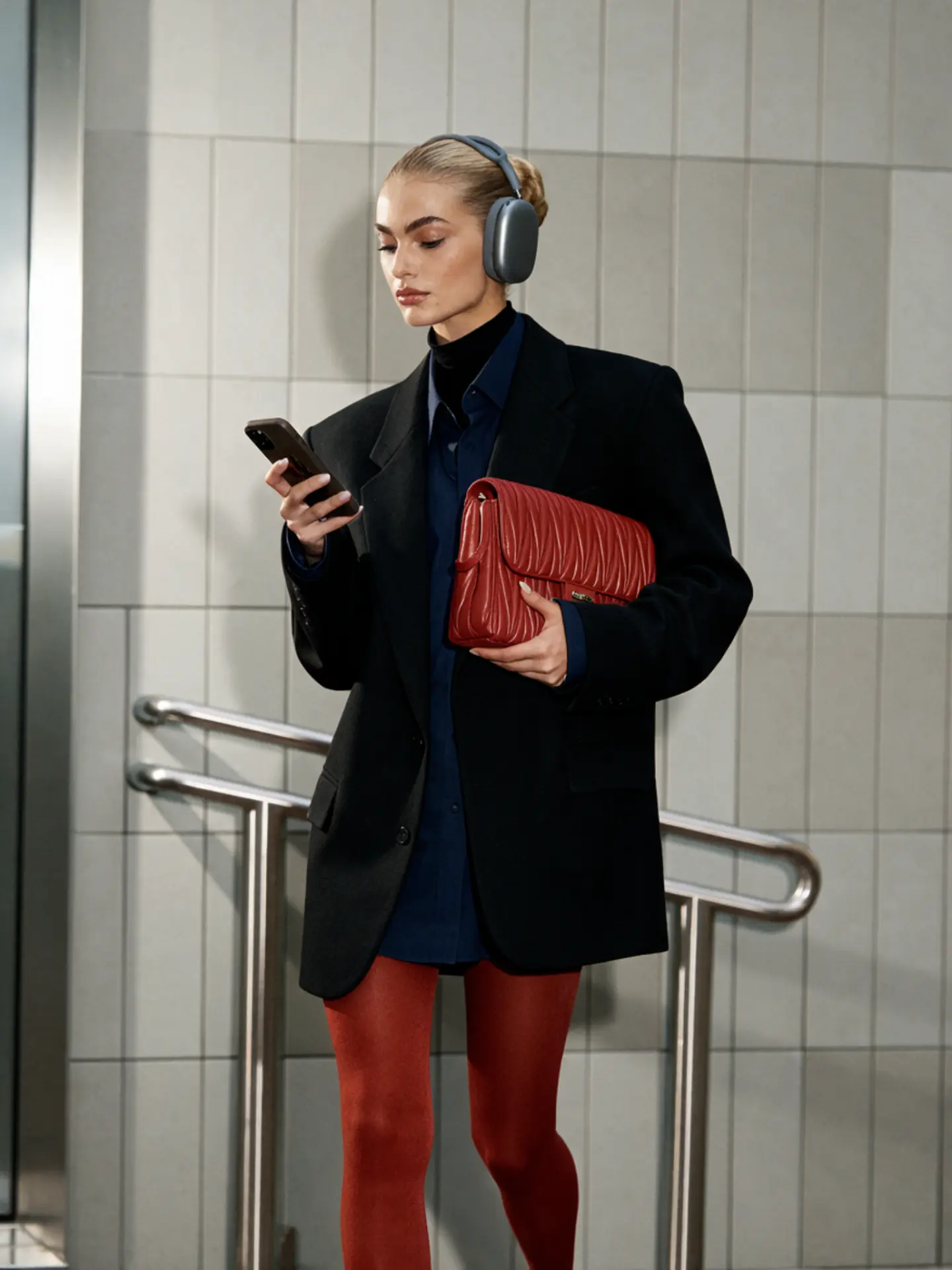 A woman in a black blazer, red tights and headphones standing by a neutral tiled wall, looking at her phone